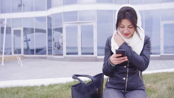 Young Brunette Woman Sits Outside with Her Phone