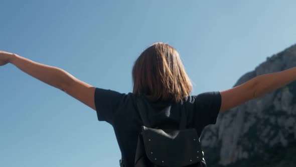 Young Woman Tourist with Backpack Standing on Cliff's Edge Top Mountain with Raised Hands and