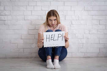 s. Frightened sad millennial woman sitting on floor and holding paper with inscription is help on white brick wall background, free space, studio shot
