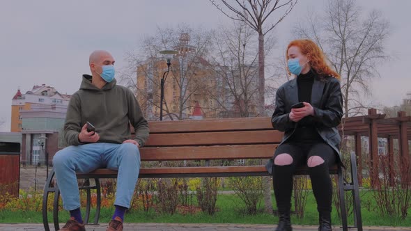 Man and Woman Sitting on Park Bench Take Off Their Protective Masks and Begin to Communicate