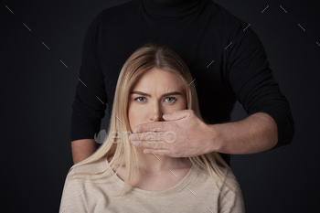 p. Male abuser in dark clothes closes mouth to sad young woman making impossible to talk, isolated on black background, free space, studio shot