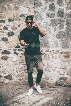 asual clothes adjusting his sunglasses and pointing you while standing against the stoned wall outdoors