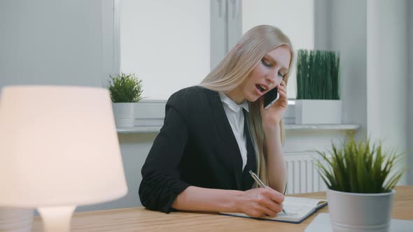 Business Woman Talking on Smartphone in Office. Elegant Young Blond Female in Office Suit Sitting at