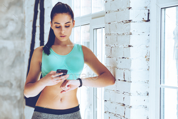 an looking at her sport bracelet and using her smartphone while leaning to the wall at gym