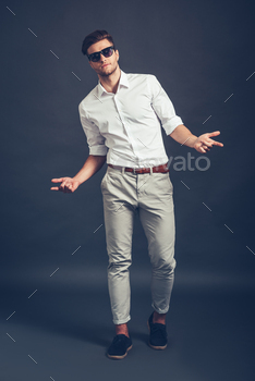 handsome man in sunglasses gesturing and looking at camera while standing against grey background
