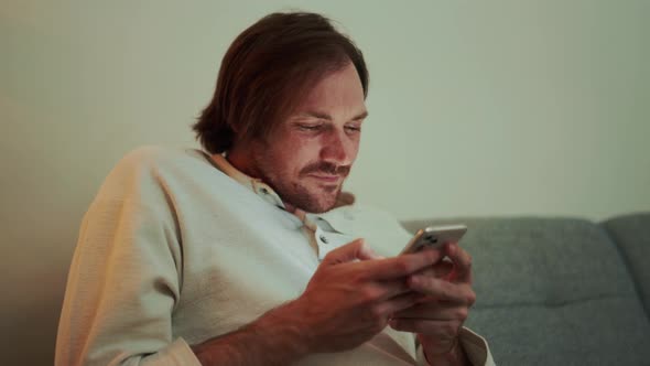 Smiling long-haired man looking at phone while sitting on the sofa