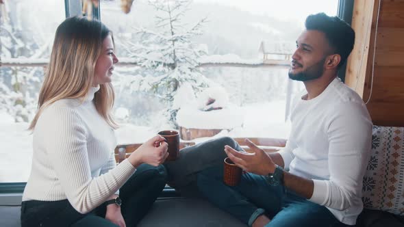 Happy Young Couple Drinking Hot Beverage and Looking at Snow Outside