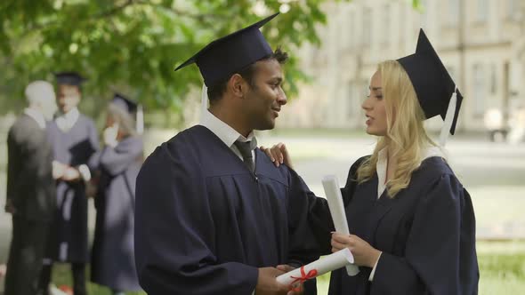 Graduates in academic regalia talking, girl fixing tie, guy kissing her on cheek