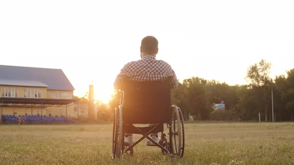 Disabled Man Sitting in Wheelchair on Field Against Sunset Raise Their Hands Up Back View