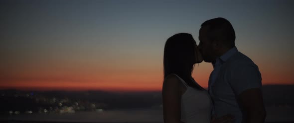 Silhouette of a couple kissing at sunset with night city lights at the background.