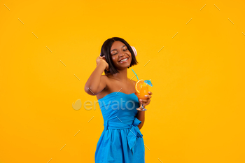Black lady in casual outfit holding tropical summer cocktail and listening to music in headphones on / Photodune, Misc