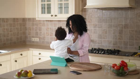 Smiling Young African American Mother Talking to Toddler Son Sitting on Countertop in Kitchen