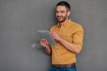 olding digital tablet and looking at it with smile while standing against grey background