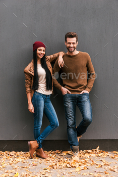 each other and smiling while leaning at the grey wall with fallen leaves laying around them