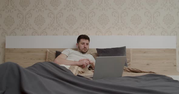 Peaceful Young Man in Pajamas Beige Tshirt Sitting in Bed Using Working on the Computer Looking Away