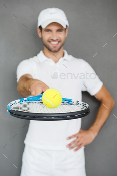 retching out tennis racket with ball and smiling while standing against grey background
