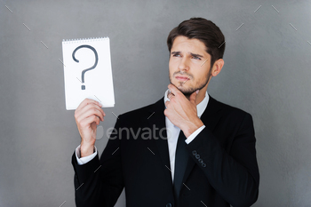 holding note pad with question mark on it and looking away while standing against grey background