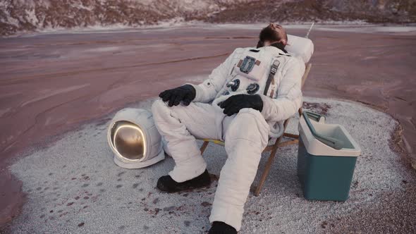 Astronaut Resting in a Chair By the Hills Without Helmet with Fridge