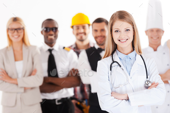  female doctor keeping arms crossed and smiling while group of people in different professions standing in the background