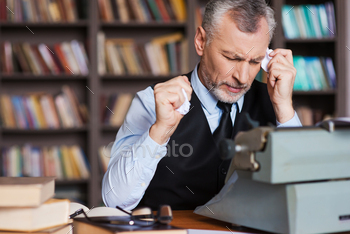 in formalwear sitting at the typewriter and holding pieces of paper in his hands with bookshelf in the background