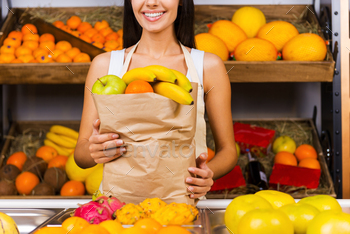 oman in apron holding paper shopping bag with fruits and smiling while standing in grocery store with variety of fruits in the background