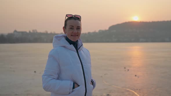 A Middleaged Girl Poses on the Shore in Winter in a White Jacket Against the Backdrop of a Beautiful