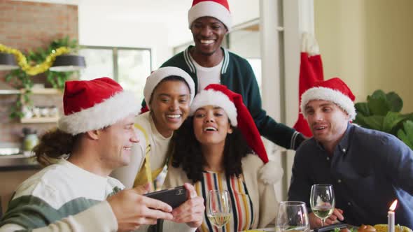 Happy group of diverse friends in santa hats celebrating meal, taking selfie at christmas time