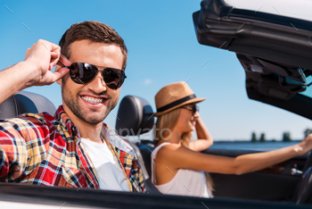 ying road trip in their convertible while handsome man adjusting his sunglasses and smiling
