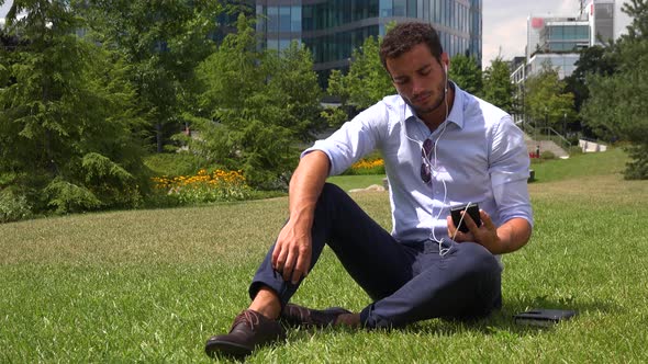 A Businessman Sits on Grass in a Park with Earphones On, Finds a Good Song on Smartphone and Listens