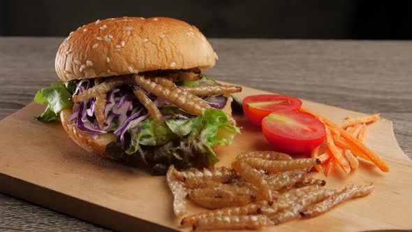 Burger with fried bamboo caterpillar and vegetable salad on wooden plate.