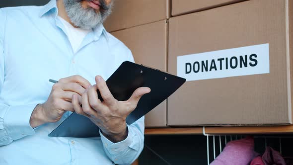 Closeup of Charity Fund Manager Writing on Clipboard Doing Inventory in Storage Room with Boxes for