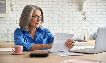 Serious middle aged woman at table holding document calculating bank loan payments, taxes, fees, retirement finances online with computer technologies