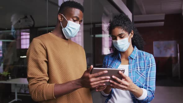 Diverse female and male business colleagues wearing masks in discussion looking at digital tablet