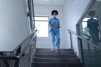 Mixed race female doctor wearing face mask and scrubs running down stairs in hospital / Photodune, Misc