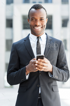 can man in formalwear holding mobile phone and smiling while standing outdoors