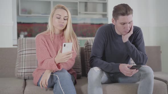 Portrait of Young Caucasian Woman and Man Sitting on Sofa and Using Smartphones. Family Members