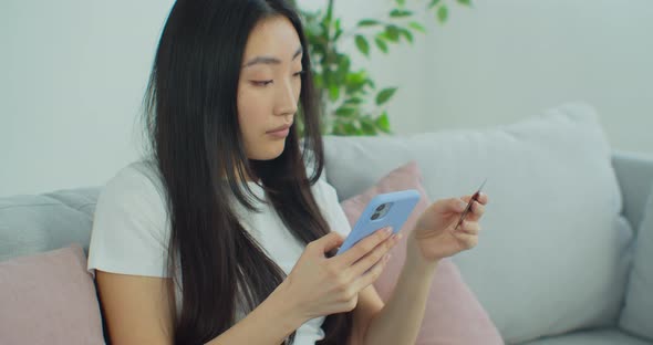 Beautiful Asian Woman Sitting on Couch Holding Cellphone Using Credit Card Makes Instant Payment