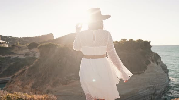 Stylish Woman in Airy White Dress Walking on Cliff Beach Near Mediterranean Sea