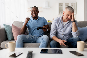 , resting at home. Sad elderly caucasian man lose, happy mature african american male rejoices to victory with joystick in living room interior