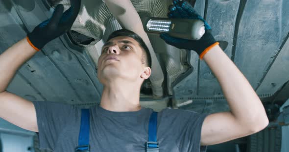 Young Man in Repair Service Examining Vehicle, Standing Under It with Light, Close Up