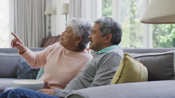 Happy biracial senior couple watching tv with popcorn and having fun at home