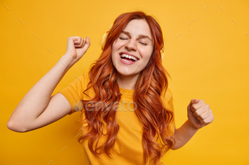 Indoor shot of happy redhead teenage girl feels relaxed shakes arms moves with rhythm of favorite mu / Photodune