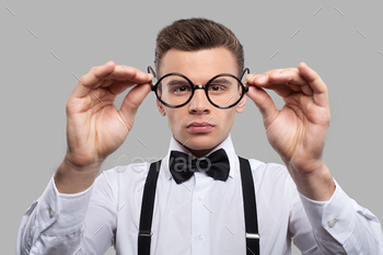 n in bow tie and suspenders holding glasses outstretched and looking through it while standing against grey background