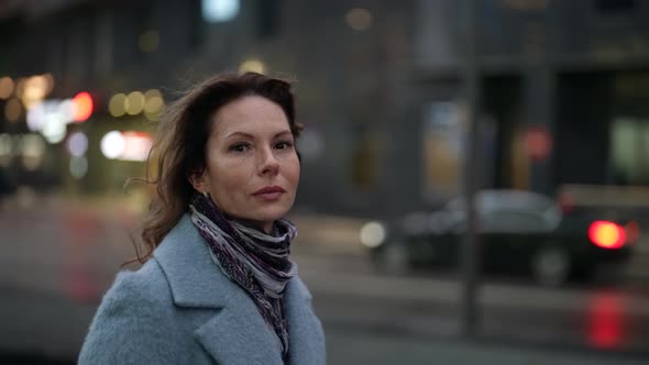 Portrait of a Woman in a Light Coat and Scarf at Dusk Against the Background of a Blurred City