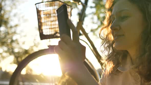 Happy Girl Looking at Phone Sitting on Grass Next To a Bicycle in the Park at Sunset, Slow Motion