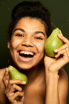  at camera, holding two juicy pears near her face, posing isolated over green background. Skincare, healthcare concept