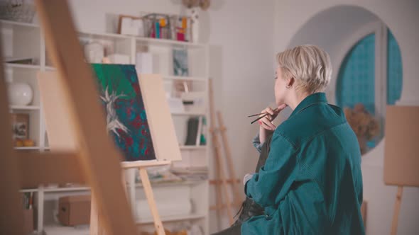 A Young Woman Artist with Short Blonde Hair Sitting in the Art Studio - Looking at Her Painting From