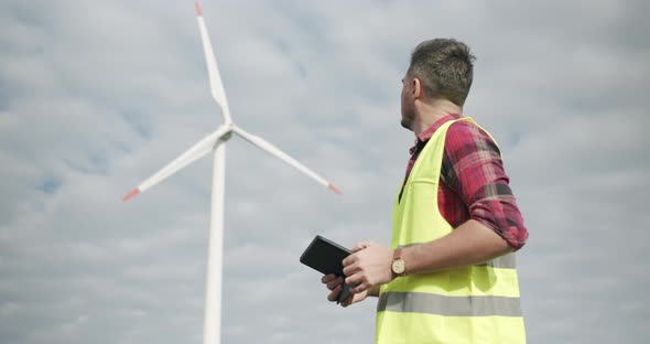 Experienced Engineer with Tablet Looks Behind on Wind Mill and Looking at Camera