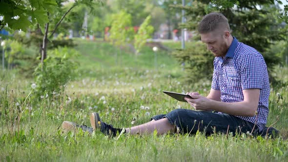 Young Caucasian Man is Swiping the Tablet Screen on the Green Lawn in City Park