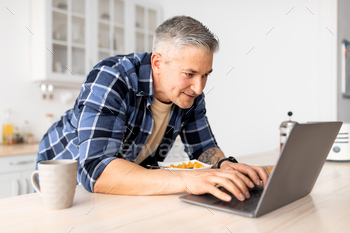 Happy aged man relaxing in kitchen with laptop and breakfast, browsing social media or reading news / Photodune, Misc